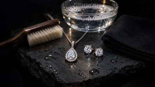 Diamond pendant necklace and diamond stud earrings on wet slate beside a soft-bristle brush, a bowl of warm soapy water, and a lint-free cloth